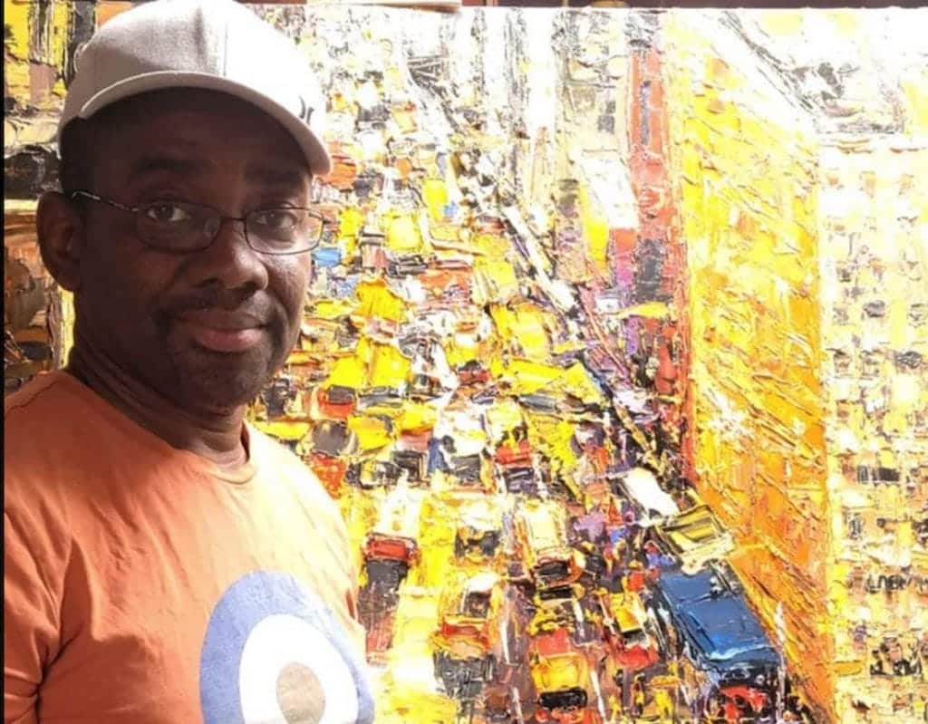 A man in a cap and glasses smiling at the camera with a colorful abstract painting in the background featuring yellow and orange tones.