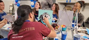 A group of people engaged in a paint and sip Washington DC activity, with one person holding up a canvas featuring a painted animal face, while others work on their artworks.