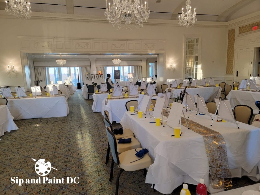 A well-lit event room is set up for a painting class with easels and seats arranged on covered tables, featuring the logo "sip and paint dc" in the corner.