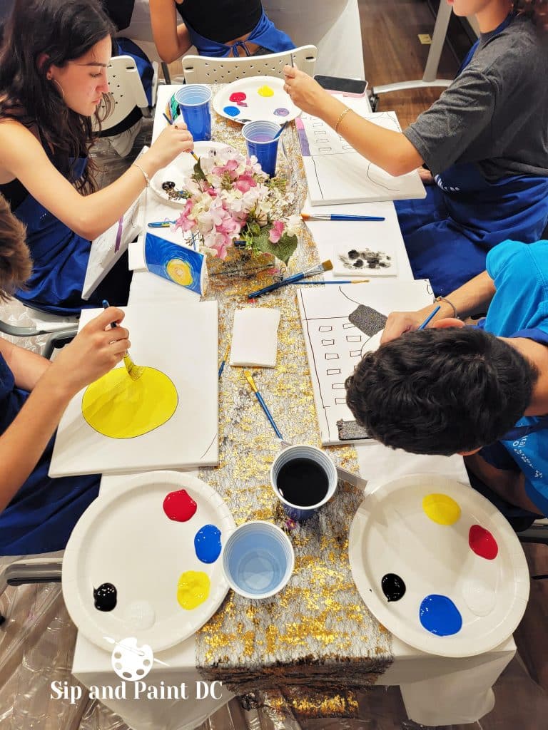 A group of people painting at a table. They each have white canvases, paint palettes with blue, red, yellow, and black paint, and blue cups. The table is decorated with flowers and a silver table runner.