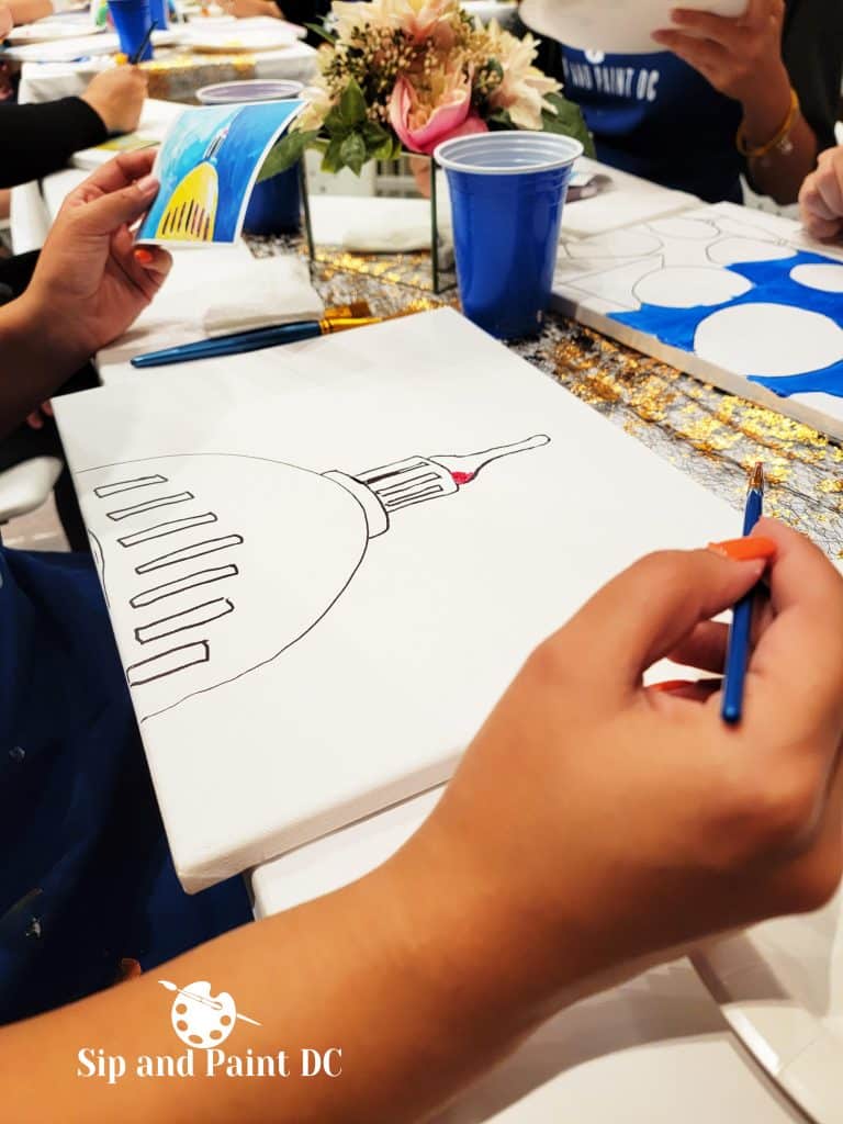 A person is painting an image of a dome on a canvas at a Sip and Paint event in Washington DC, with paint supplies and a reference photo visible.