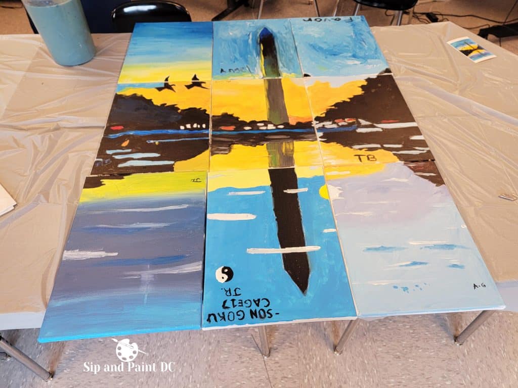 Three painted canvases laid out on a table, depicting a split landscape of birds flying over a sunset, water reflecting the scene, and the Washington Monument. The setup is from an event by Sip and Paint DC.