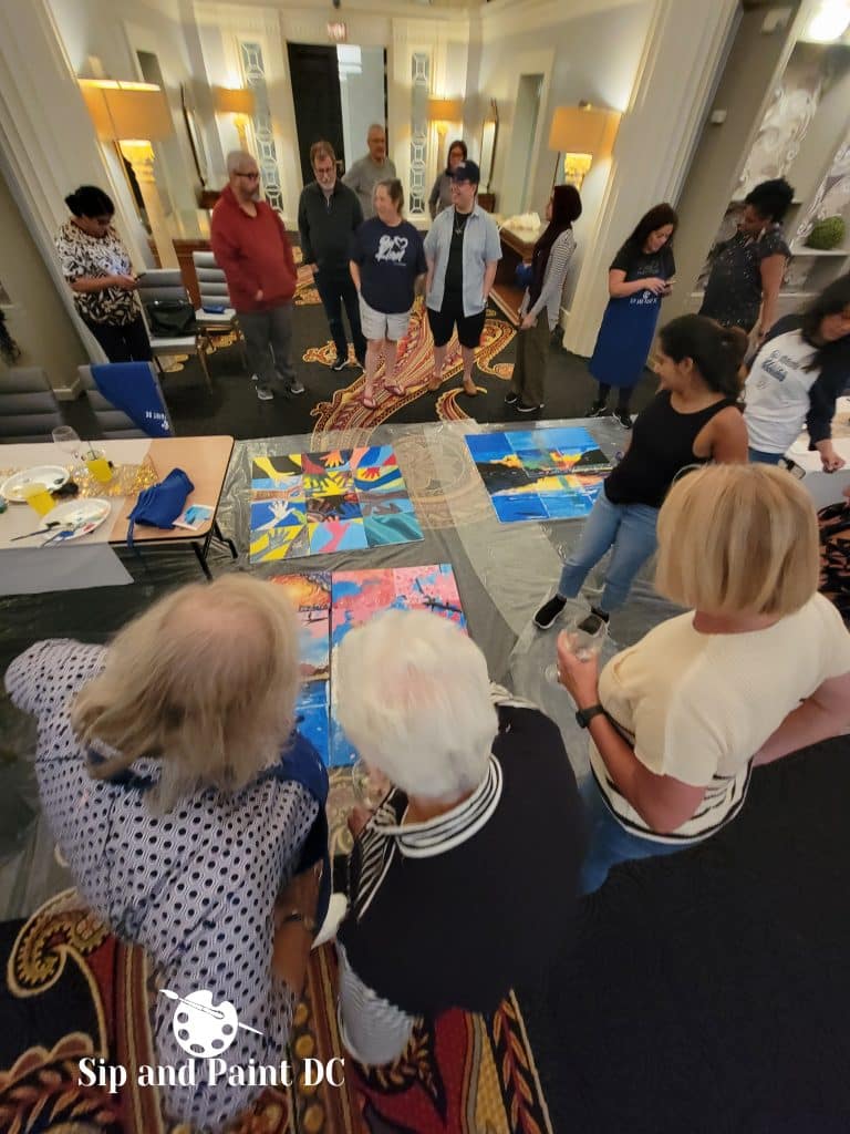 A group of people at an art event stand around paintings on the floor, observing and discussing the artwork. The event setting appears to be indoors with various lighting fixtures.
