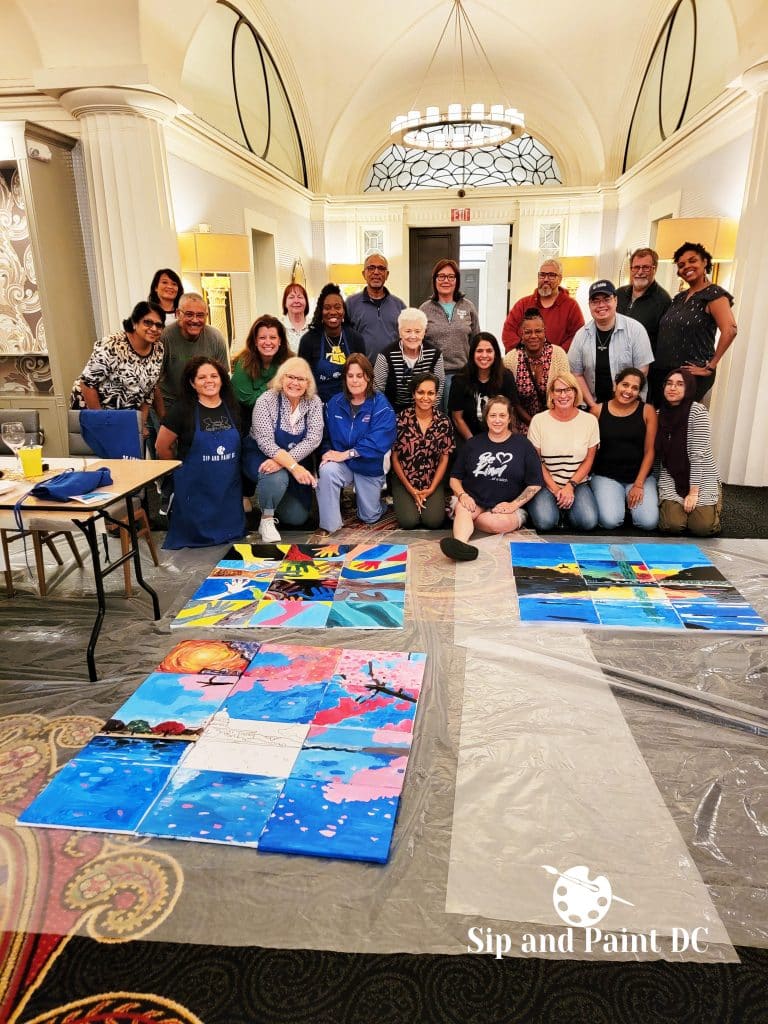 A group of people posing together indoors at a painting event called "Sip and Paint DC." Several completed canvases are displayed on the floor in front of them.