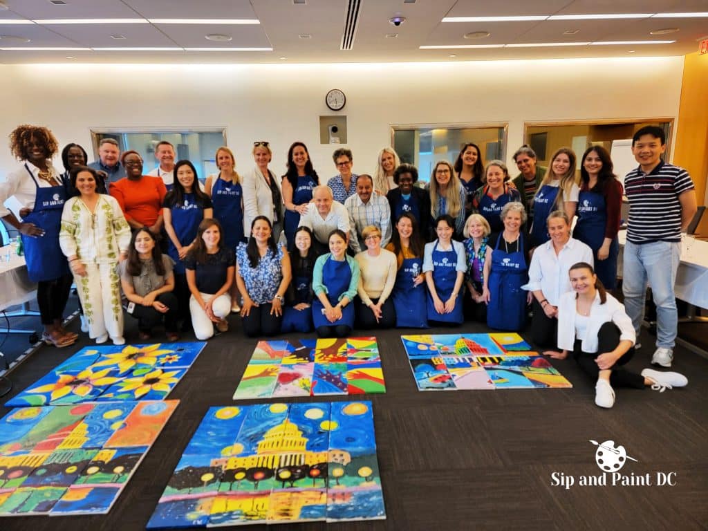 A group of people posing with colorful paintings on the floor at an indoor art event. The paintings depict various vibrant scenes. The room features white walls and a clock above a doorway.