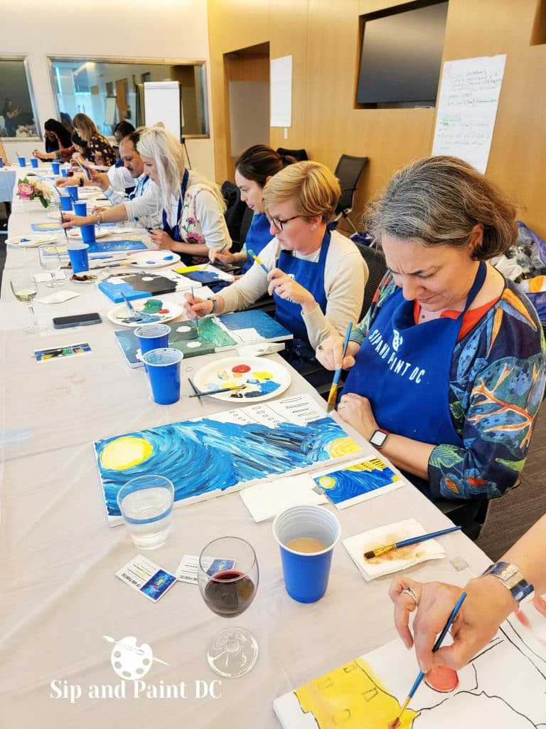 A group of people sits at a long table, each working on their own painting. They are wearing aprons and focusing on their artwork. Brushes, paint, and cups are on the table. A "Sip and Paint DC" logo is visible.