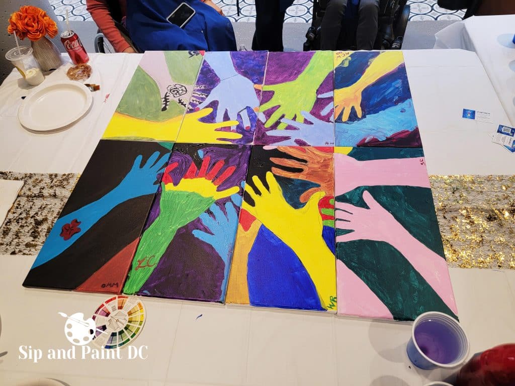 A set of colorful paintings arranged to form an image of multiple hands reaching towards the center, displayed on a table with art supplies around. The tablecloth has "Sip and Paint DC" branding.
