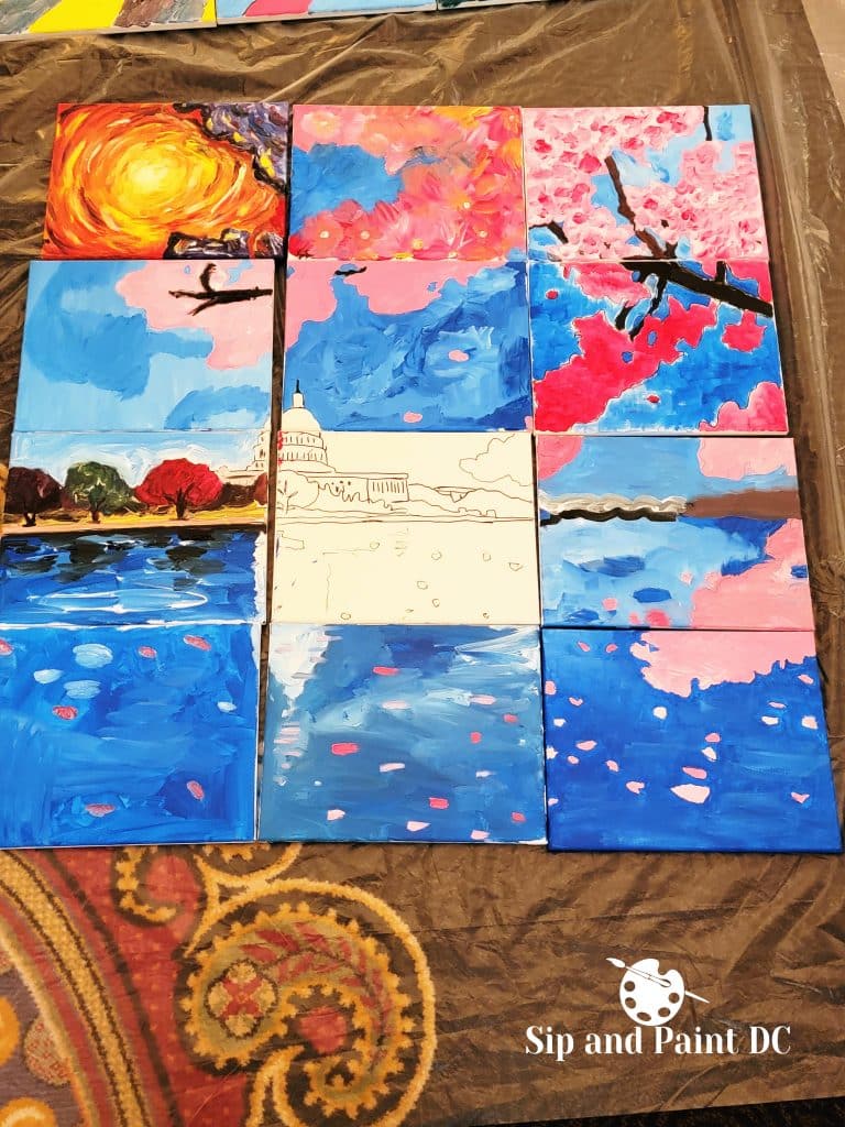 A collection of nine canvases displaying colorful paintings, including landscapes, cherry blossoms, a cityscape, and abstract art, arranged on a brown tablecloth with a "Sip and Paint DC" logo.