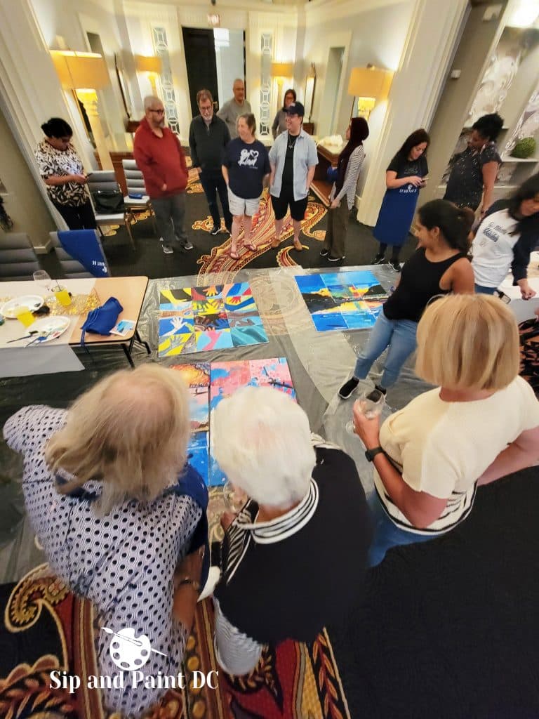 A group of people at a "Sip and Paint DC" event, standing around and viewing colorful paintings laid out on the floor in a well-lit room.