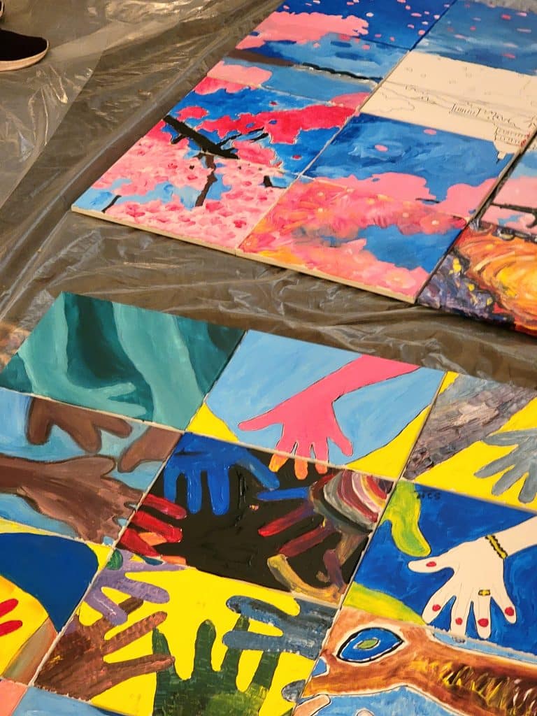 Multiple colorful painted tiles featuring hands and various abstract designs are arranged on a surface covered with clear plastic, reminiscent of a lively sip and paint session.