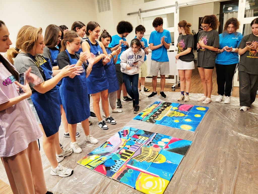 A group of people in a room clapping around two colorful paintings placed on the floor. Some wear paint-stained aprons.