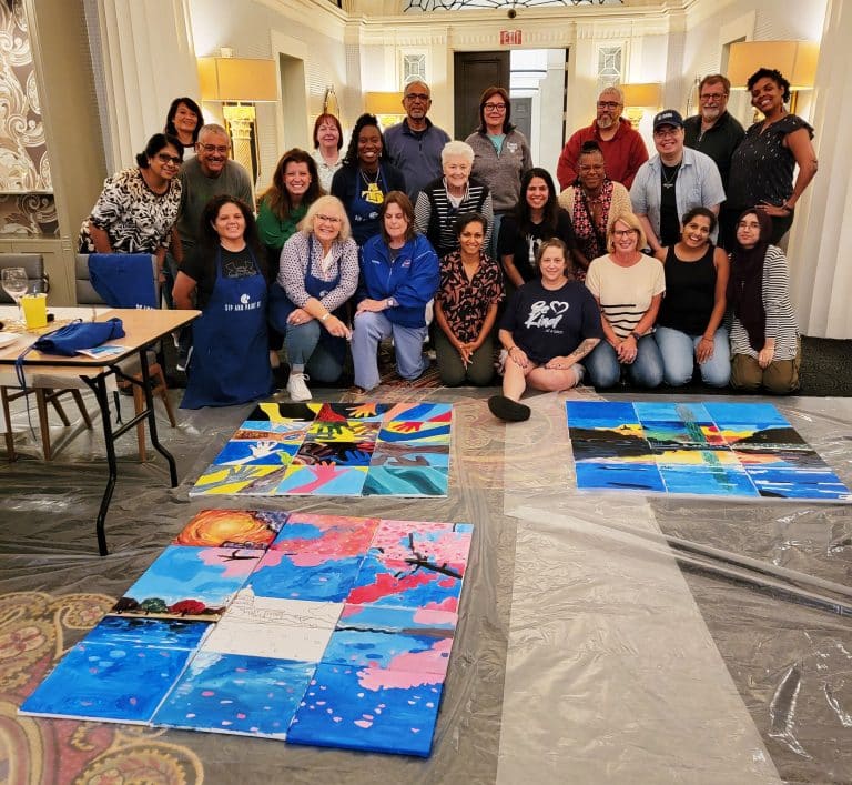 A group of people pose indoors around colorful paintings on the floor. They are in a decorated room with ornate details and lighting.