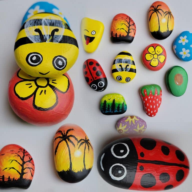 Colorful stones painted with various designs: bees, a ladybug, flowers, landscapes, and a strawberry. The stones are arranged on a white background.