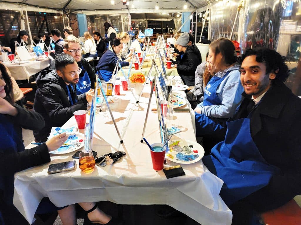People seated at long tables enjoy a lively paint and sip event, wearing blue aprons as they create art on canvases. Art supplies, drinks, and food fill the tables under a canopy, making for a fun and creative gathering.