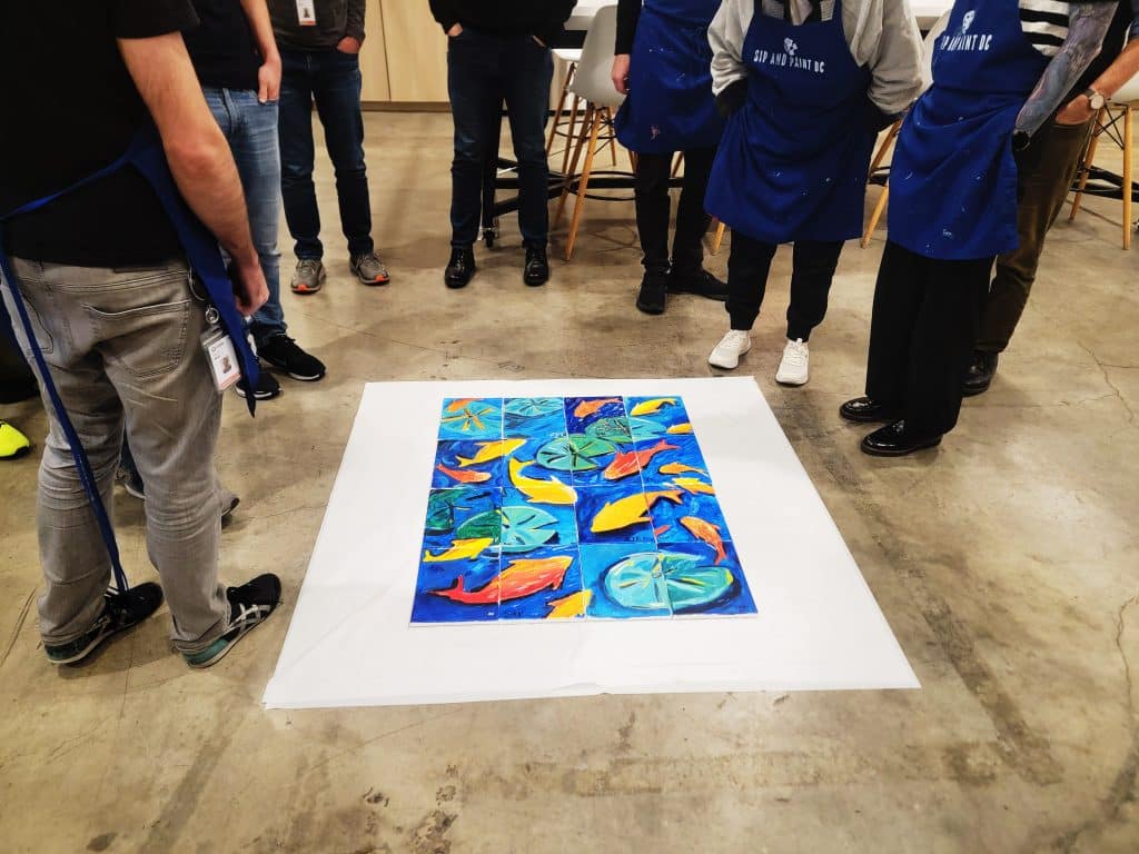 A group of people in aprons stand in a circle on a concrete floor, looking at a colorful abstract painting laid out on white paper.